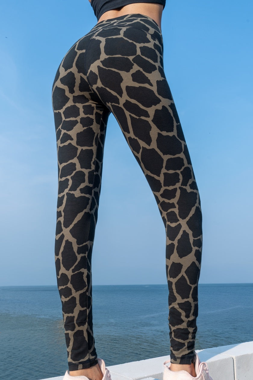 Woman wearing Funky Simplicity Giraffe Leggings in the colour Olive Black shown from the back with an ocean and blue sky on the background