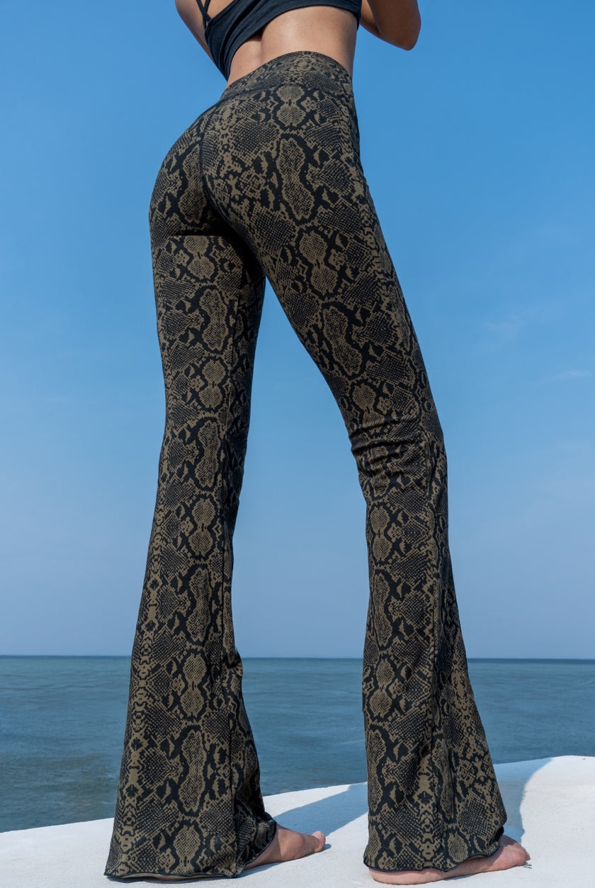 Woman wearing olive black Snake patterned flared leggings shown from the back, from Funky Simplicity - standing on a white surface with a blue sky and ocean background