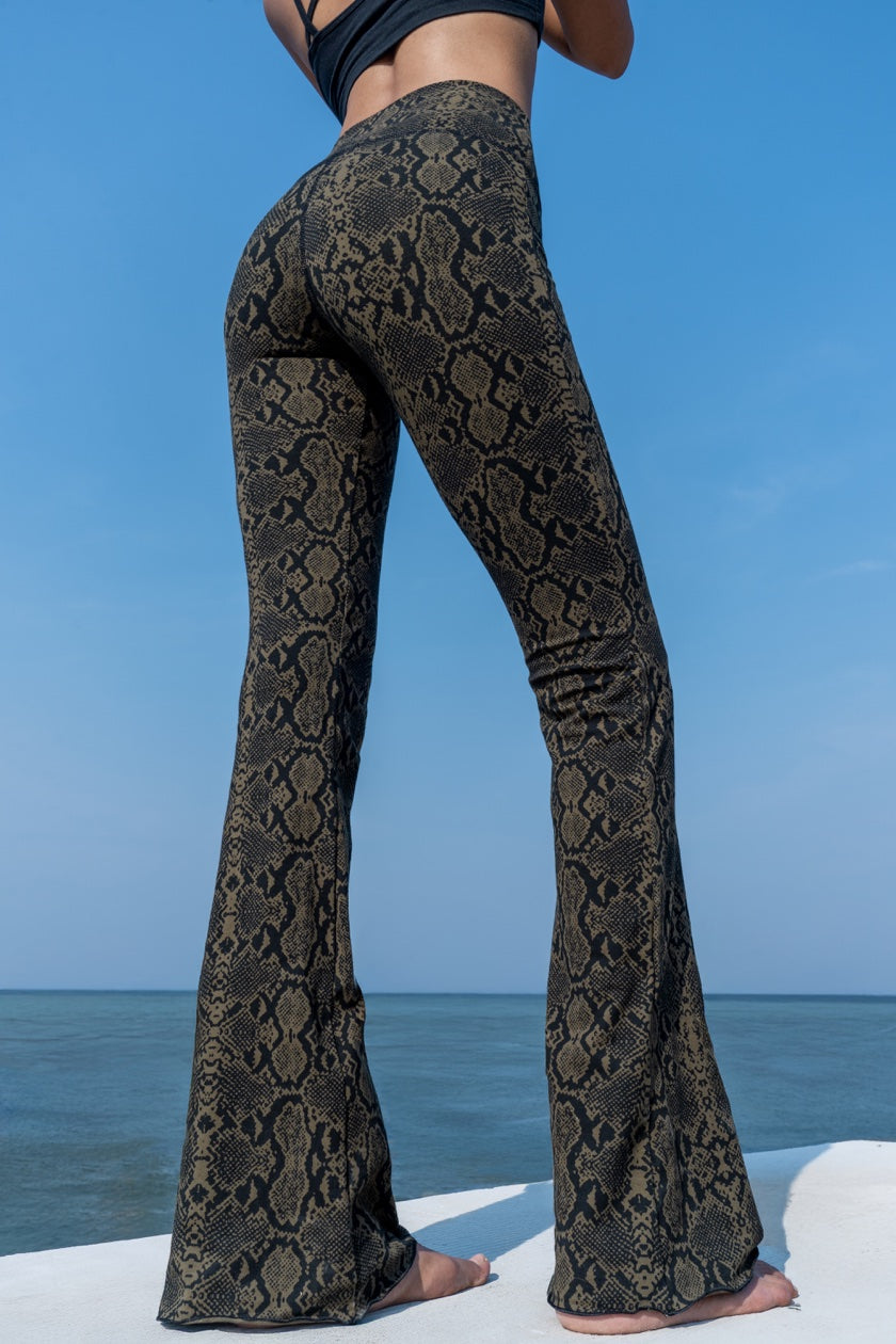 Woman wearing olive black Snake patterned  flared leggings shown from the back, from Funky Simplicity - standing on a white surface with a blue sky and ocean background